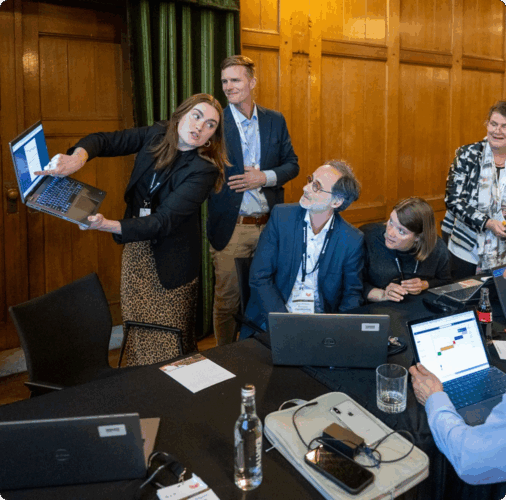 A Boeing employee is standing holding a laptop and pointing to the screen during a workshop on Cascade. Around them are several seated attendees watching the demonstration.