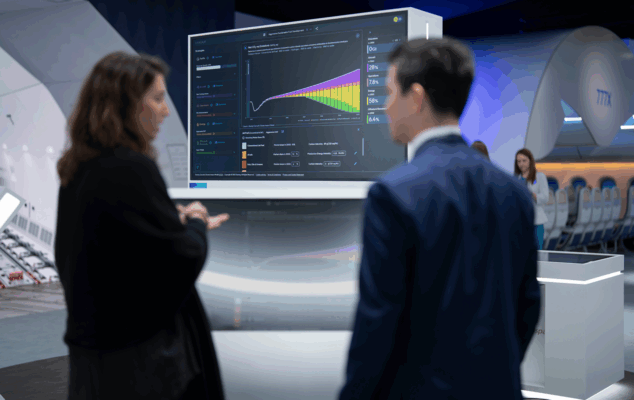 Two people are having a conversation in front of a large screen at an aviation conference. The screen displays the Boeing Cascade app.
