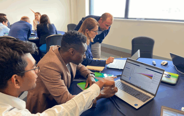A workshop on using Cascade. Attendees are sat in groups at different tables looking at Cascade on laptops. A Boeing employee stands at the shoulder of one of the attendees.